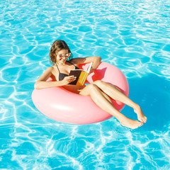 bikini girl with sunglasses relaxed and reading book on pink inflatable pool ring