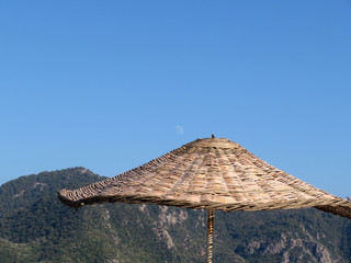 Sun umbrella with a wicker roof on the background of mountains and blue sky. Sun awning, wicker parasol. Tan and beach vacation concept