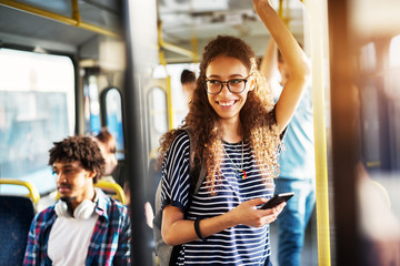 Young gorgeous cheerful woman is standing on the bus using the phone and smiling. - obrazy, fototapety, plakaty