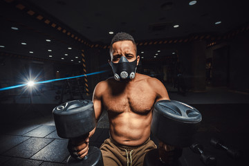 Fototapeta na wymiar african american athletic man in mask sitting and waiting and looking at camera before lifting dumbbells in front of the mirror. black man in gym with dumbbells close-up