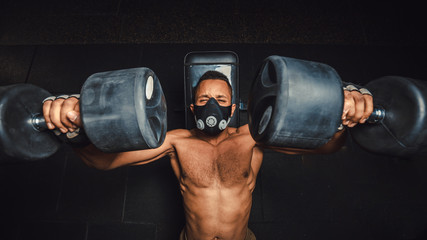 Fototapeta na wymiar african american athletic man in mask lifting dumbbells and working on his chest view from above. black man in gym doing exercise for chest with dumbbells close-up