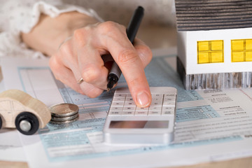 Young woman makes some calculations. Small paper house, coins, car in the background.