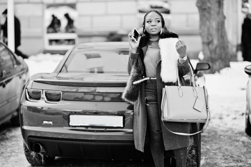 Rich african american girl in red coat and fur against red muscle car speaking on mobile phone....