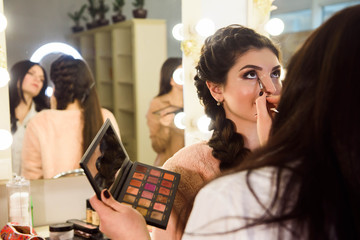 Make-up artist applying bright base color eyeshadow on model's eye and holding a shell with eyeshadow on background, close up