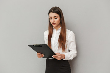 Fototapeta premium Photo of serious woman in white shirt and black skirt holding clipboard with documents in office, isolated over gray background