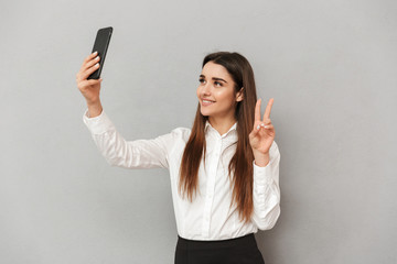 Fototapeta premium Gorgeous brunette woman in business wear taking selfie on cell phone and gesturing victory sign on camera, isolated over gray background
