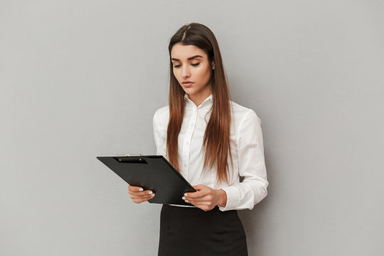 Photo of serious woman in white shirt and black skirt holding clipboard with documents in office, isolated over gray background