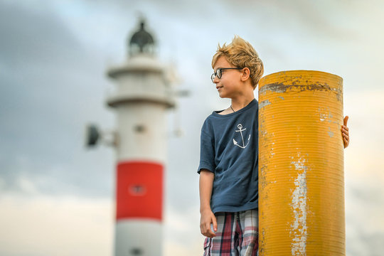 Boy with sun glasses standing in front of a lighthouse in Tenerife, looking at the horizon. Wearing a blue t-shirt with an anchor, enjoying a beautiful sunset after a fun day at the beach. 