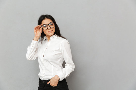 Portrait of smiling successful woman with long brown hair wearing office clothing looking aside and touching her eyeglasses, isolated over gray background