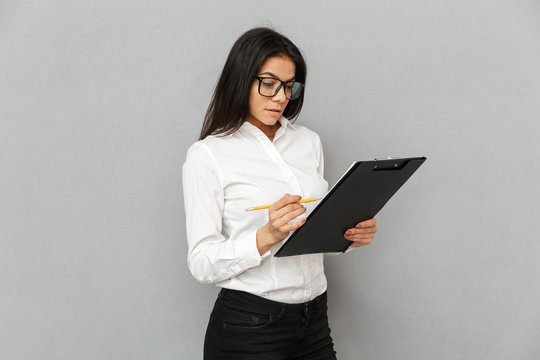 Portrait of successful woman wearing businesslike outfit and glasses holding clipboard with papers and writing down notes in documents, isolated over gray background