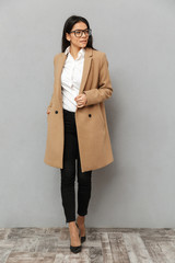 Full length image of fashion woman in glasses and beige coat walking on heels and looking aside, isolated over gray background
