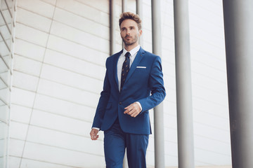 Handsome young man model standing by the glassy office building wearing a blue classic tuxedo suit.