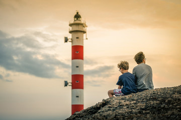 Two young boys with glasses sitting on the rocks looking the sunset. A teenager and his son with...