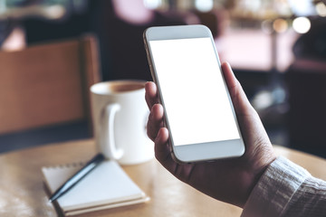 Mockup image of hand holding white mobile phone with blank desktop screen with coffee cup and notebook in cafe
