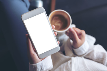 Top view mockup image of woman's hands holding white mobile phone with blank screen while drinking coffee in cafe