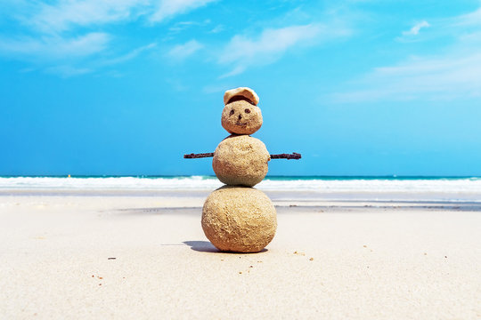 Christmas positive Sandy Snowman in red Santa Claus hat at ocean sunset beach.