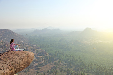 A young bald man sits on top of a mountain against a background of sunrise or sunset and looks...