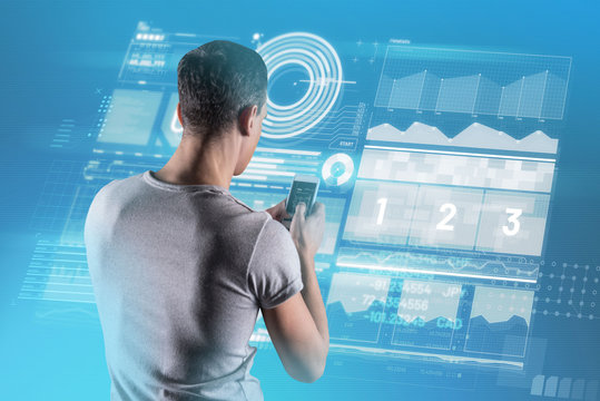 Useful device. Calm attentive analytics manager holding a modern gadget while standing in front of a transparent screen