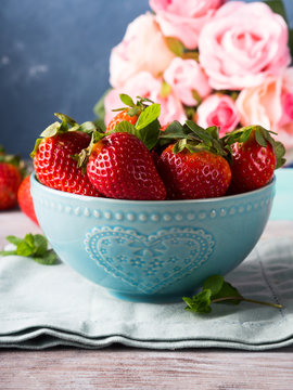Fresh spring strawberries in blue bowl with heart and pink roses in the background