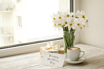 White yellow daffodil, narcissus flowers in glass vase on wooden windowsill, no window wiev. Happy mother's day text greeting card and craft paper prsent. Close up, copy space, still life, background.
