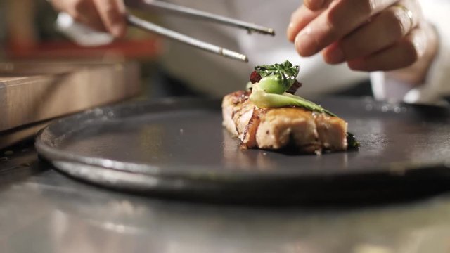 Hands chef finish off the dish. Slow motion. Chef at the white uniform to spread on large plate piece of meat, vegetables and seasonings, prepares to serve the client a restaurant, cafe or catering