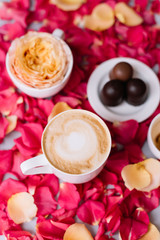 Fototapeta na wymiar Delicious fresh morning cappuccino coffee with a thick milk foam, some chocolate sweets and a blooming flower in a cup on the pink rose petals background