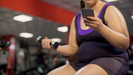 Overweight lady using fitness application on gadget, doing dumbbell exercise