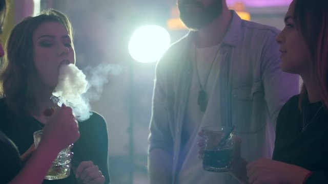 young sexy woman enjoys hookah and exhale smoke near friends who holds cocktails at night party