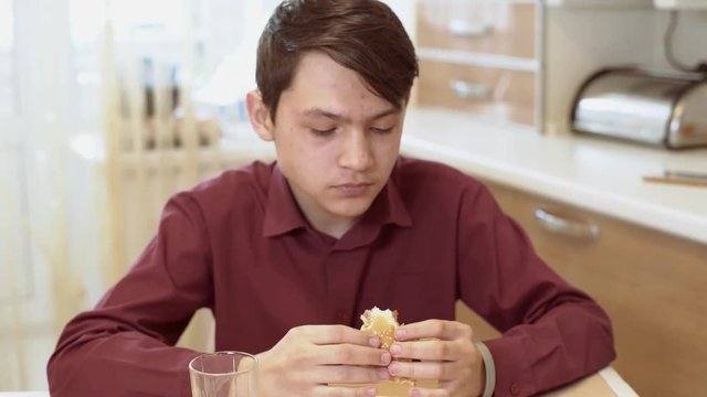 A 15 year old teen bites a sandwich. A young guy eats a sandwich at a table.