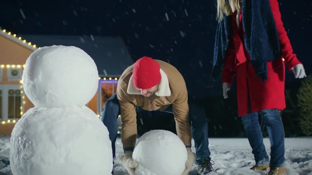 Happy Young Family Making Snowman in the Backyard of their Idyllic House. Father Rolls Snowball and Puts it on top of the Other, Daughter and Wife Help Him. 