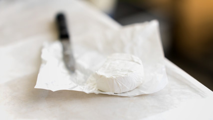 Small head of brie cheese in unfolded paper packaging, knife. Gastronomic dainty products on market counter, real scene in the food market. Selectiv focus, for background