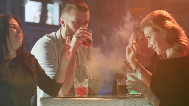 young friends having fun near table with colorful beverages in smoke on background of colored lamps at night party