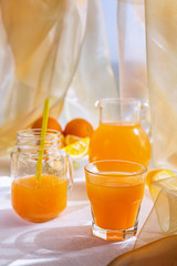 Juicy orange juice in a glass and jug and ripe fruit orange on a white table near the window, the concept of breakfast on a sunny summer day