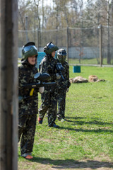 Boy in the camouflage holds a paintball gun  in one hand and protective helmet , standing on the field with group of players on the background