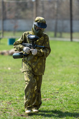 Boy in the camouflage holds a paintball gun  in one hand and protective helmet , standing on the field with group of players on the background
