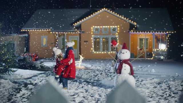 On the Happy Winter Evening Young Father, Mother and Cute Little Daughter Play in Snowballs, Running around the Snowman They've Built in the Backyard of Their Idyllic House Decorated with Garlands. 