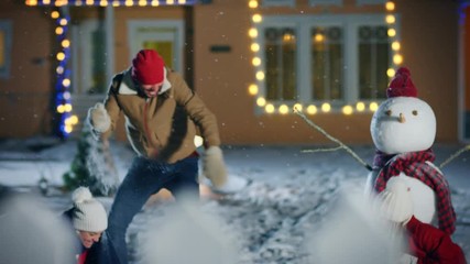 On the Happy Winter Evening Young Father, Mother and Cute Little Daughter Play in Snowballs, Running around the Snowman They've Built in the Backyard of Their Idyllic House Decorated with Garlands. - Powered by Adobe
