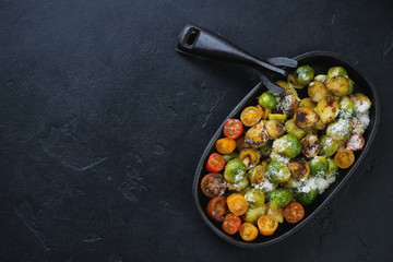 Cast-iron pan with fried brussels sprouts, cherry tomatoes and grated cheese. View from above on a black stone background, horizontal shot, copy space