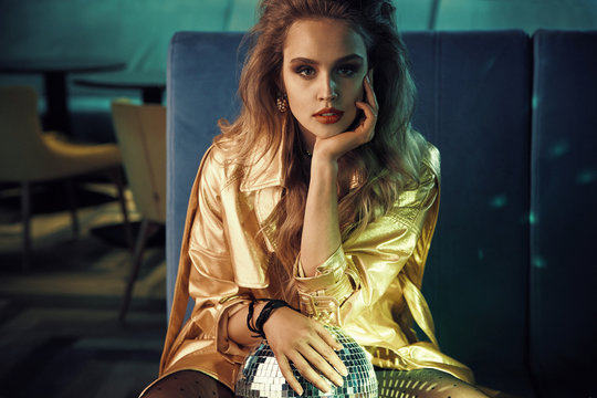 Luxury portrait of beautiful young woman in gold trench coat, sitting on blue couch with disco ball. Retro style