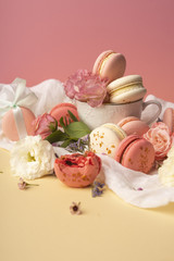 Pink and white macaroons cakes with big and small flower buds are decoratively laid on a white fabric and a cup on a yellow background