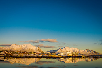 Sunset at Henningsvaer shoreline with factory and huge mountains covered with snow in a gorgeous blue sky reflecting in the lake on Lofoten Islands, Austvagoya