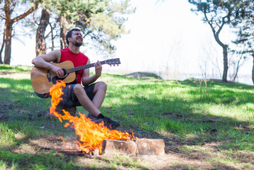 Young man playing acoustic guitar while sitting near campfire.