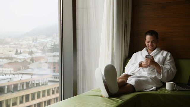 Young smiling handsome man wearing bathrobe messaging on a mobile phone and smiling. He is sitting near window with great city view with cup of coffee at luxury scyscraper hotel.