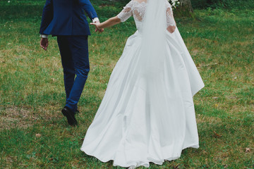 Wedding couple are holding hands in the green forest. Groom and bride in dress with train and tulle veil are walking outdoors. High green trees and bright emerald green grass.