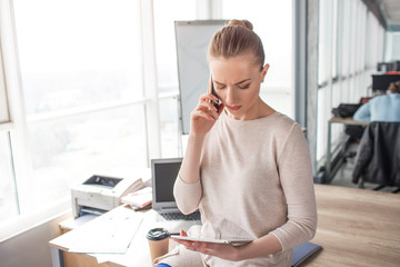 Business woman is looking at the tablet and talking on the phone at the same time. She has a phone meeting with client. This woman stands in a big, large room near window.