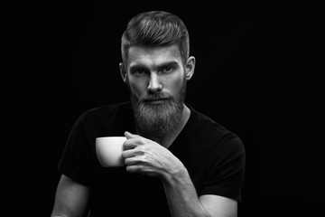 Black and white dramatic light studio shot of bearded stylish young man with cup of coffee in hand Handsome man having coffee look at camera Male taking rest and drinking fresh morning espresso . - obrazy, fototapety, plakaty