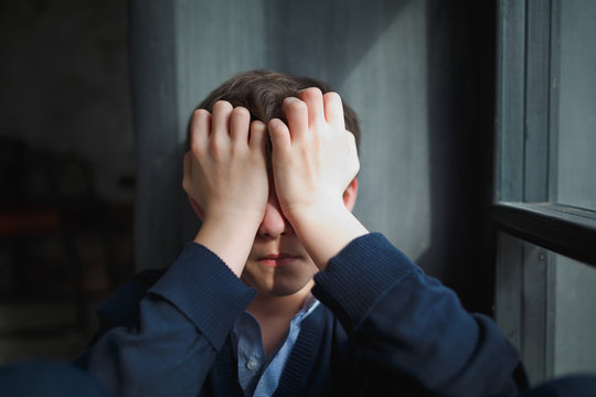 Pensive Sad Teenager Boy With Blue Shirt And Blue Jeans Sits At The Window And Closes His Face With His Hands. The Sun's Rays Fall On The Boy's Face.