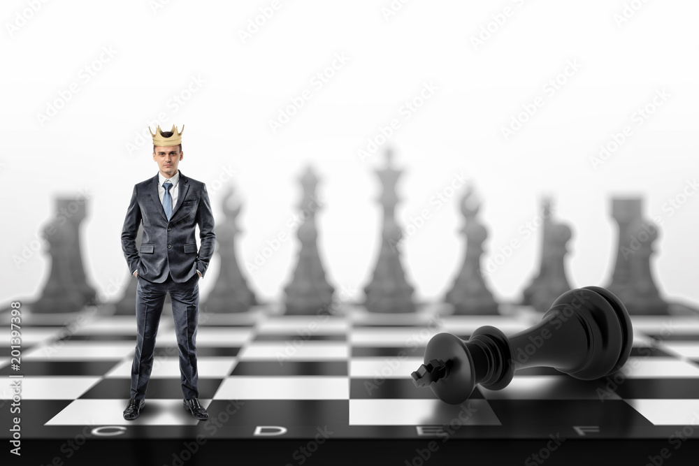 Wall mural A small businessman with a golden crown on his head stands on a chessboard near a fallen black king.