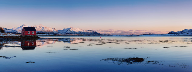 Landscape with beautiful winter lake, red rorbu house and snowy mountains at sunset at Lofoten...
