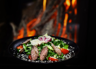 Dish of beef or veal served with lettuce leaves, tomatoes and sprinkled with parmesan cheese, fire on background. Delicious meat served in restaurant on black dishes. Restaurant dish concept.